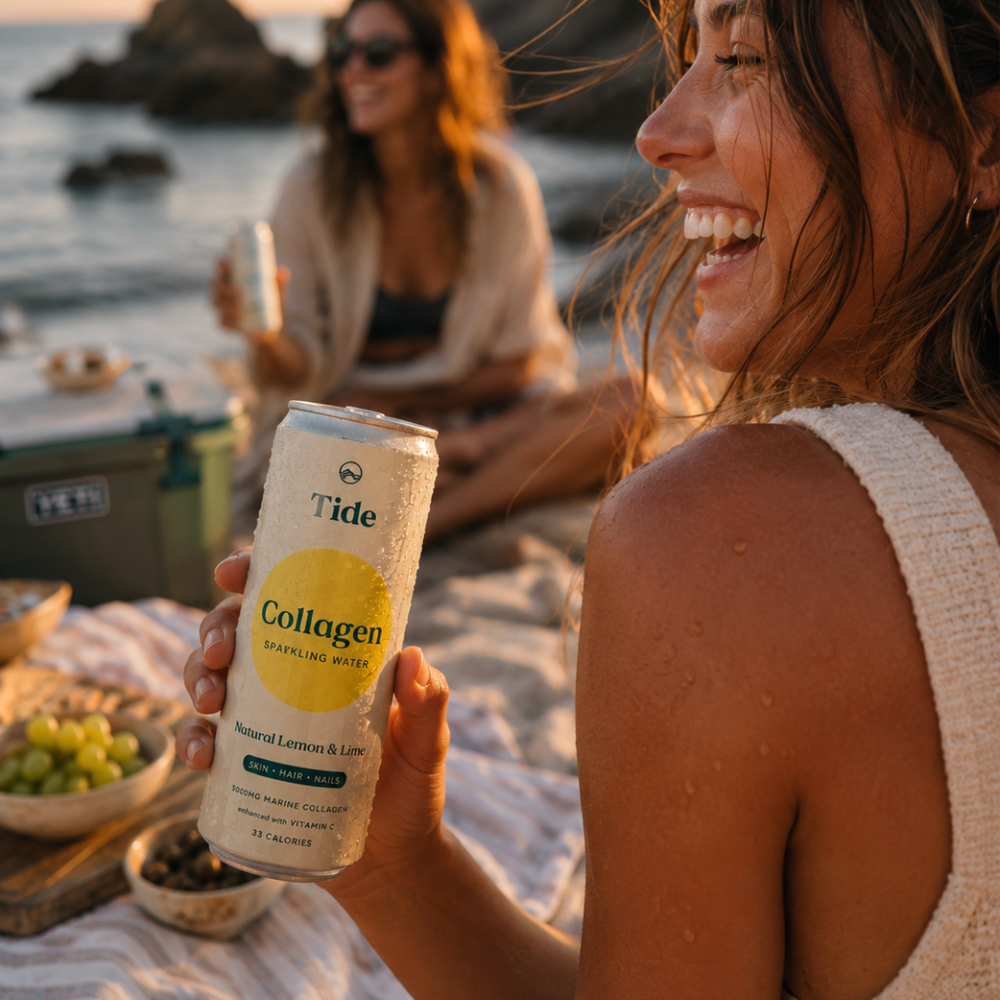 A woman on a beach picnic holds a can of Tide Collagen sparkling water. The can is white with a yellow circle and black text, and the background shows the ocean, rocks, and another woman in the distance.