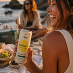 A woman on a beach picnic holds a can of Tide Collagen sparkling water. The can is white with a yellow circle and black text, and the background shows the ocean, rocks, and another woman in the distance.