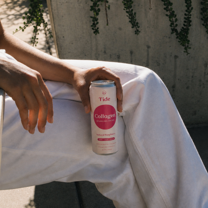 A person in white clothing holds a can of Tide Collagen drink mix outdoors against a concrete wall with hanging greenery.