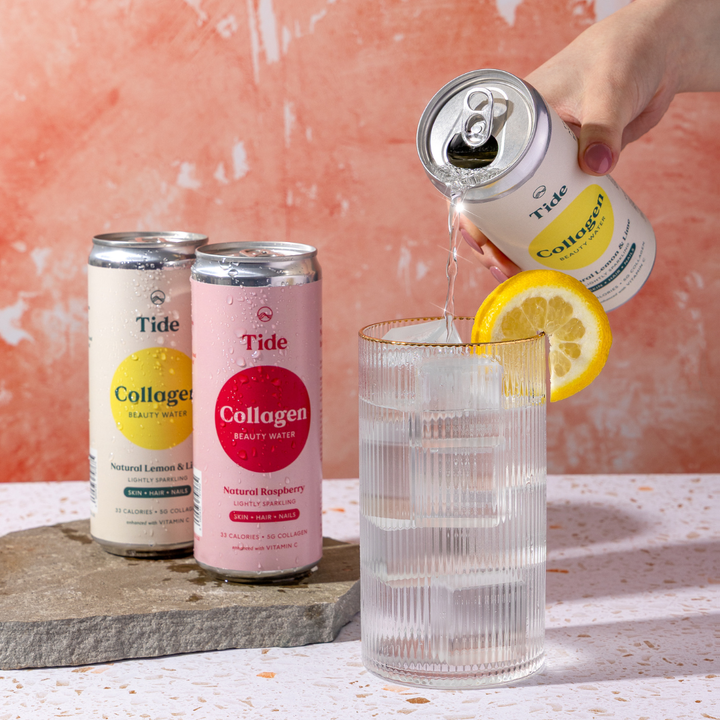A hand pours Tide Collagen Beauty Water from a silver can into a clear ribbed glass with a lemon slice garnish. Two more Tide Collagen cans, one cream with a yellow circle and one pink with a red circle, stand on a stone slab against a peach textured background.