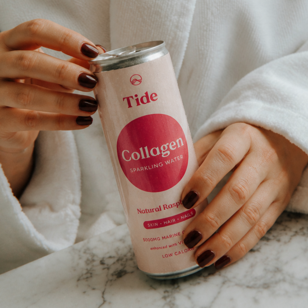 A person in a white robe holds a can of Tide Collagen Sparkling Water with dark brown manicured nails against a marble surface.