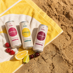 Three Tide Collagen beverage cans arranged on a yellow-and-white towel on sandy ground, with raspberries, a lemon slice, and a lime nearby. The cans show flavors: Collagen Lemon & Lime, Collagen Sparkling Cherry, and Collagen Wild Berry Raspberry.