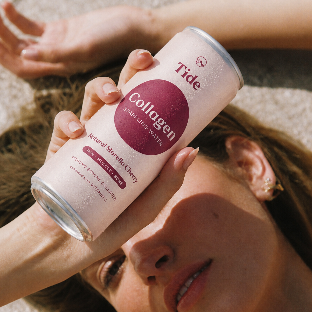 A person lying on sand holds a can of Tide Collagen sparkling water. The can is pale pink with a large magenta circle label and text reading 'Tide Collagen Natural Morello Cherry.